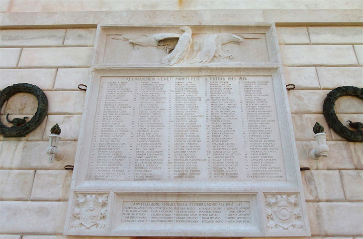 Dogana da Mar, lapide commemorativa Lastra con iscrizione a niello definita da cornice modanata; in alto, comparto rettangolare scolpito a rilievo, raffigurante un gruppo di cinque aquile. Al centro, epigrafe dedicata ai caduti della grande guerra; e sotto, altra iscrizione in memoria dei soldati deceduti durante il secondo conflitto mondiale. In basso, coppia di comparti quadrati angolari, scolpiti a rilievo, e raffiguranti il leone marciano "in moleca" a sinistra, e lo stemma della Guardia di Finanza a destra, entrambi entro scudo affiancato da fronde di quercia e di alloro.